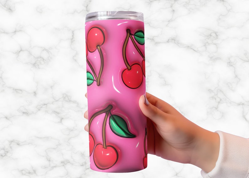 May include: A pink tumbler with a repeating pattern of red cherries with green leaves. The cherries appear to be 3D, with a glossy, raised effect. The tumbler is cylindrical and has a silver lid.