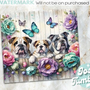 Op de afbeelding: Drie bulldog puppy's kijken over een houten hek versierd met kleurrijke bloemen en vlinders. Het hek is wit geschilderd met een houtnerfstructuur. De bloemen zijn roze, paars, blauw en groen. De vlinders zijn blauw en paars.