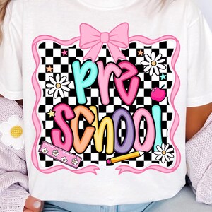 May include: White t-shirt with a colorful "Pre School" graphic. The design features a pink bow, daisies, stars, a ruler, a pencil, and an apple against a black and white checkered background. The text is in various bright colors.