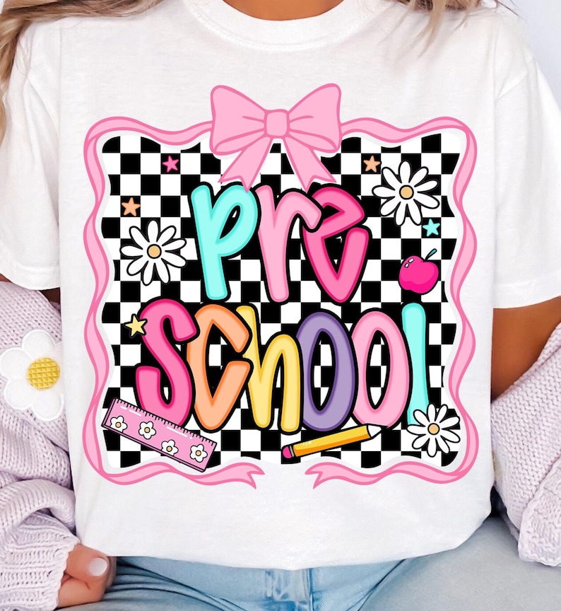 May include: White t-shirt with a colorful "Pre School" graphic. The design features a pink bow, daisies, stars, a ruler, a pencil, and an apple against a black and white checkered background. The text is in various bright colors.