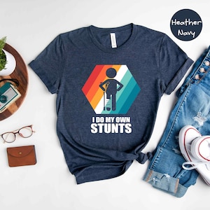 May include: A heather navy blue t-shirt with a retro-style graphic of a person with a broken leg and crutches. The text "I do my own stunts" is printed below the graphic.