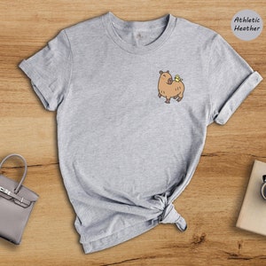 May include: Gray t-shirt with a cartoon capybara wearing a yellow bird on its back. The t-shirt is labeled "Athletic Heather".