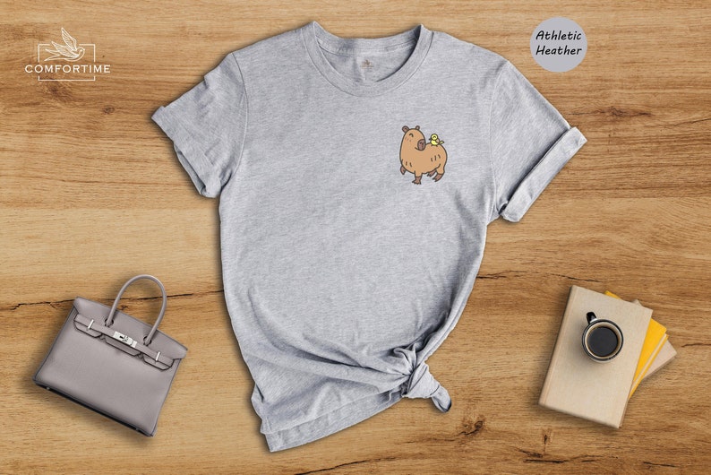 May include: Gray t-shirt with a cartoon capybara wearing a yellow bird on its back. The t-shirt is labeled "Athletic Heather".
