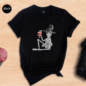 Pu&ograve; includere: Maglietta nera con una grafica di uno scheletro che indossa un cappello da strega e tiene un bicchiere di liquido rosso. Lo scheletro &egrave; seduto con le gambe incrociate.