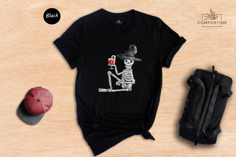 Pu&ograve; includere: Maglietta nera con una grafica di uno scheletro che indossa un cappello da strega e tiene un bicchiere di liquido rosso. Lo scheletro &egrave; seduto con le gambe incrociate.