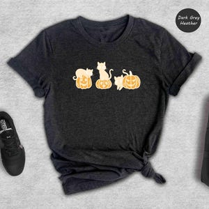 May include: Dark grey heather t-shirt featuring a graphic of three white cats sitting on top of orange pumpkins.