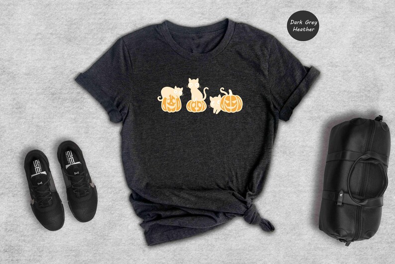 May include: Dark grey heather t-shirt featuring a graphic of three white cats sitting on top of orange pumpkins.