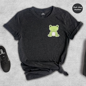 Op de afbeelding: Donkergrijs heather t-shirt met een cartoon kikker afbeelding. De kikker is groen met een witte buik en lacht. Het shirt ligt plat op een grijs oppervlak.