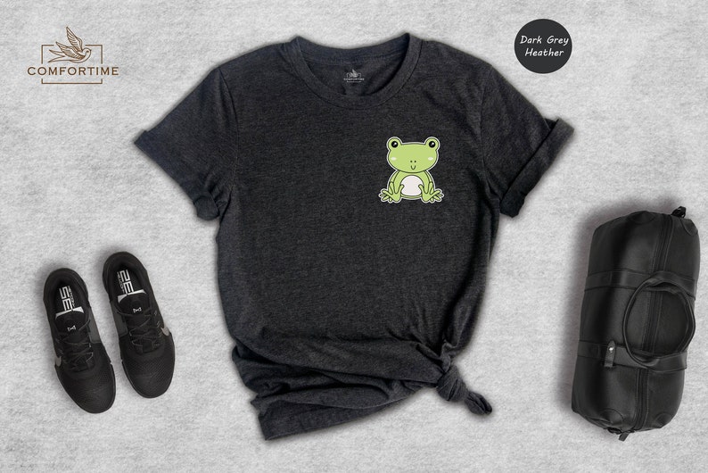 Op de afbeelding: Donkergrijs heather t-shirt met een cartoon kikker afbeelding. De kikker is groen met een witte buik en lacht. Het shirt ligt plat op een grijs oppervlak.