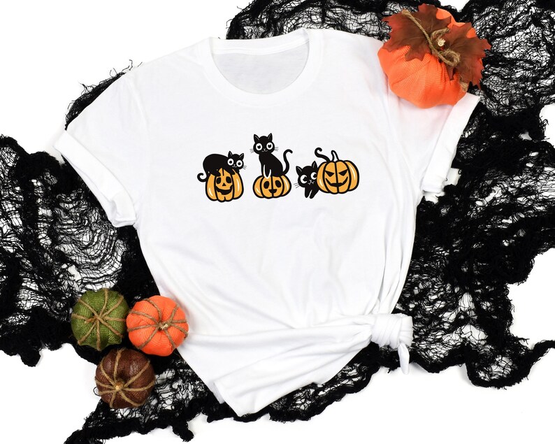 May include: A white t-shirt with a graphic of three black cats sitting on top of orange pumpkins with jack-o-lantern faces. The pumpkins are outlined in black. The t-shirt is displayed on a black lace fabric background.