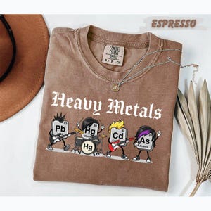 Peut inclure: Un t-shirt marron avec le texte Heavy Metals et un graphique représentant quatre personnages de dessin animé symbolisant les éléments chimiques Pb, Hg, Cd et As jouant des instruments. Le t-shirt a un col rond et une étiquette Comfort Colors.