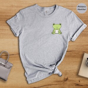 Op de afbeelding: Grijs T-shirt met een groene kikker afbeelding op de voorkant. Het shirt is gemaakt van athletic heather stof.