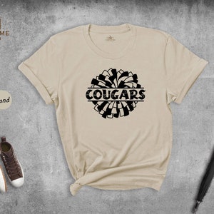 May include: A sand colored t-shirt with a black and white pom-pom design and the word "COUGARS" printed in black.