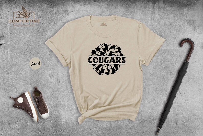 May include: A sand colored t-shirt with a black and white pom-pom design and the word "COUGARS" printed in black.