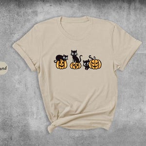 May include: A beige t-shirt featuring a design of three black cats perched on top of jack-o'-lantern pumpkins.
