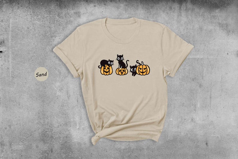 May include: A beige t-shirt featuring a design of three black cats perched on top of jack-o'-lantern pumpkins.
