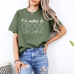 May include: A woman wearing a green t-shirt with the text "I'd rather be cross stitching" in white. She is wearing blue jeans with distressed details.