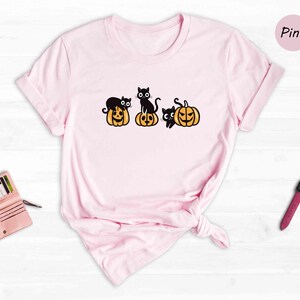 May include: A pink t-shirt featuring a design of three black cats perched on pumpkins. The pumpkins are orange with black faces. The word "Pink" is in the top right corner of the image.
