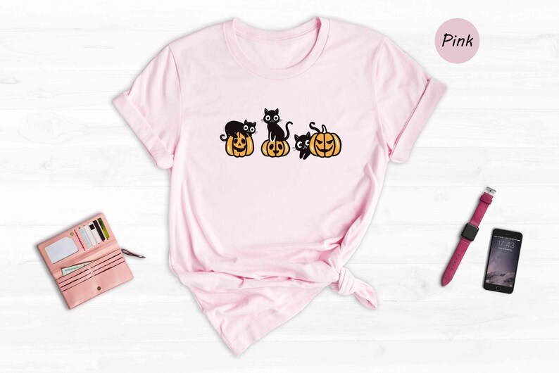 May include: A pink t-shirt featuring a design of three black cats perched on pumpkins. The pumpkins are orange with black faces. The word "Pink" is in the top right corner of the image.
