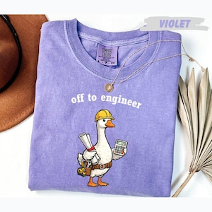 Peut inclure: Un t-shirt violet avec un motif représentant une oie blanche portant un casque de chantier jaune, tenant un plan roulé et une calculatrice, avec une ceinture à outils. Le texte "off to engineer" est imprimé en blanc au-dessus du motif.