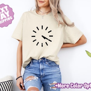 May include: A beige t-shirt with a black clock graphic. The clock has a distressed look and the hands are pointing to 12 o'clock.