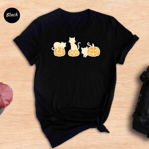 May include: Black t-shirt featuring a design of three cats sitting on pumpkins. The cats are white with orange markings. The pumpkins are orange with black jack-o'-lantern faces.