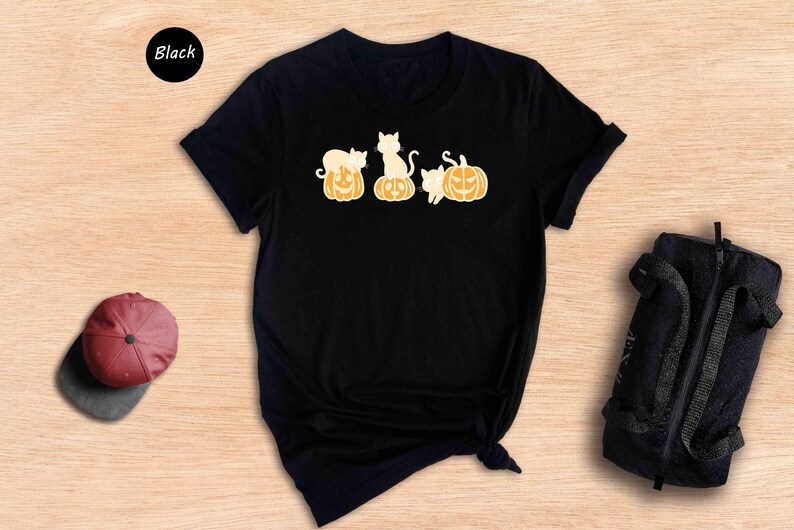 May include: Black t-shirt featuring a design of three cats sitting on pumpkins. The cats are white with orange markings. The pumpkins are orange with black jack-o'-lantern faces.