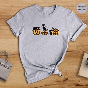 May include: A grey t-shirt featuring a black cat and pumpkin design. The text "Athletic Heather" is in the top right corner of the image.