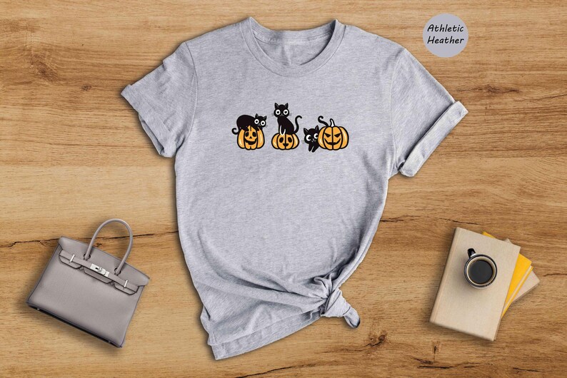 May include: A grey t-shirt featuring a black cat and pumpkin design. The text "Athletic Heather" is in the top right corner of the image.