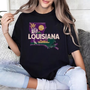May include: A black t-shirt with a purple and green outline of the state of Louisiana. The outline includes a small boat, a pelican, and palm trees. The text "LOUISIANA" is printed in white on the shirt.