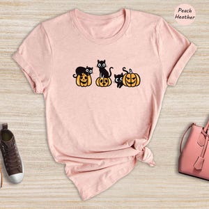 May include: A peach heather t-shirt featuring a graphic of black cats sitting on pumpkins. The graphic depicts four cats and four pumpkins.