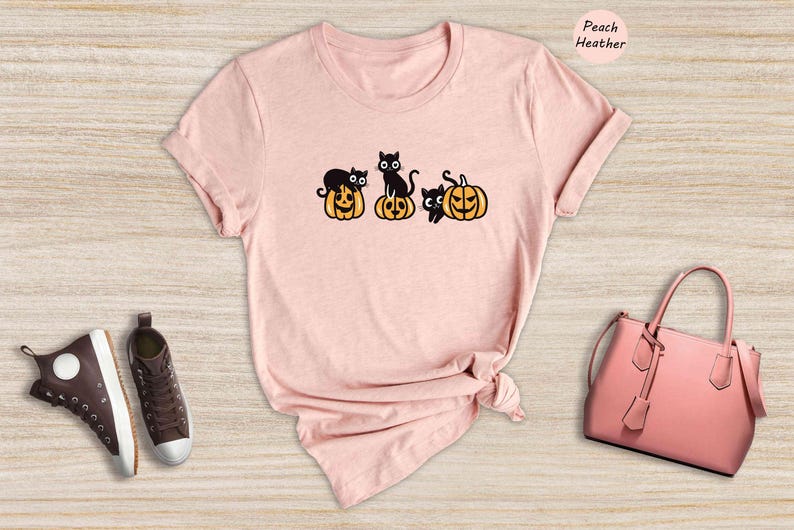 May include: A peach heather t-shirt featuring a graphic of black cats sitting on pumpkins. The graphic depicts four cats and four pumpkins.