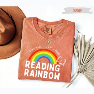 May include: A terracotta-colored t-shirt featuring a graphic of a rainbow and an open book. The text reads, "Take a Look, it's in a Book!" and "READING RAINBOW" in white capital letters. A thin gold-tone necklace rests on the shirt.
