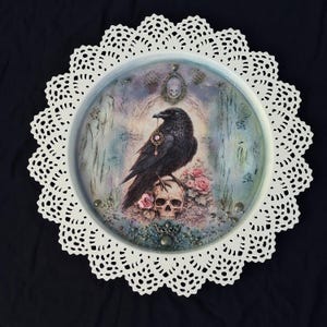 May include: A round decorative tray with a black raven perched on a skull, surrounded by pink roses. The tray has a white lace doily border. The raven wears a pendant. The background is a misty scene. The tray's diameter is approximately 30 cm.