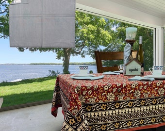May include: A red and black floral tablecloth with a white border, set for a dinner party with plates, glasses, and a centerpiece. The table is set in a dining room with a view of a lake and trees.