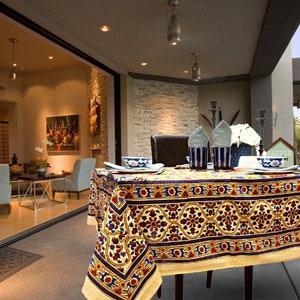 May include: A yellow tablecloth with a blue, red, and gold floral pattern. The tablecloth is laid on a table with four chairs and a bowl on each place setting. The table is set on a patio with a stone wall in the background.