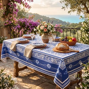 May include: A rectangular dining table covered with a blue and white floral patterned tablecloth. The table is set with white plates, beige napkins, clear glasses, a ceramic vase with flowers, a wooden board with bread, and a bowl of fruit.