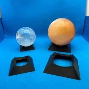 May include: Four black, 3D printed, square-shaped display stands for crystal balls. Two stands are holding a clear crystal ball and a light orange crystal ball.