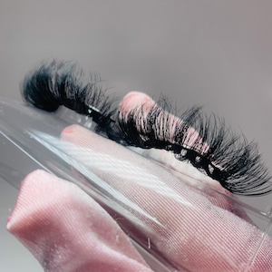 May include: Two pairs of black false eyelashes in a clear plastic container. The container is on a pink fabric surface.