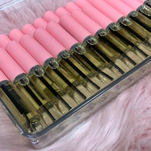 K&ouml;nnte beinhalten: Ein transparenter Plastikbeh&auml;lter gef&uuml;llt mit mehreren rosa und goldenen Flaschen mit Wimpernserum. Die Flaschen haben einen schwarzen Applikatorpinsel.