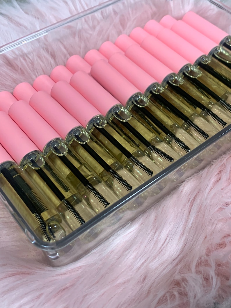 K&ouml;nnte beinhalten: Ein transparenter Plastikbeh&auml;lter gef&uuml;llt mit mehreren rosa und goldenen Flaschen mit Wimpernserum. Die Flaschen haben einen schwarzen Applikatorpinsel.
