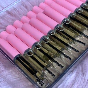 K&ouml;nnte beinhalten: Ein transparenter Plastikbeh&auml;lter gef&uuml;llt mit mehreren rosa und durchsichtigen R&ouml;hrchen, die eine klare Fl&uuml;ssigkeit mit einem schwarzen Applikatorpinsel enthalten. Die R&ouml;hrchen sind in Reihen angeordnet.