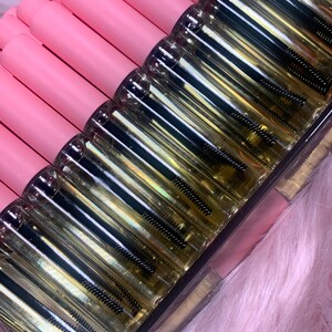 K&ouml;nnte beinhalten: Nahaufnahme einer durchsichtigen Glasflasche mit einem schwarzen Applikatorpinsel. Die Flasche ist mit einer klaren Fl&uuml;ssigkeit gef&uuml;llt und hat einen rosa Deckel. Die Flaschen sind in einer Reihe auf einer rosa Oberfl&auml;che angeordnet.