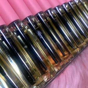 K&ouml;nnte beinhalten: Nahaufnahme von mehreren durchsichtigen Glasflaschen mit schwarzen Applikatorst&auml;bchen. Die Flaschen sind mit einer hellgelben Fl&uuml;ssigkeit gef&uuml;llt und in Reihen auf einer rosa Oberfl&auml;che angeordnet.