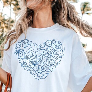 May include: White t-shirt featuring a blue heart-shaped design filled with nautical elements. The design includes seashells, starfish, a sailboat, and a palm tree. The shirt is a casual, everyday wear item.