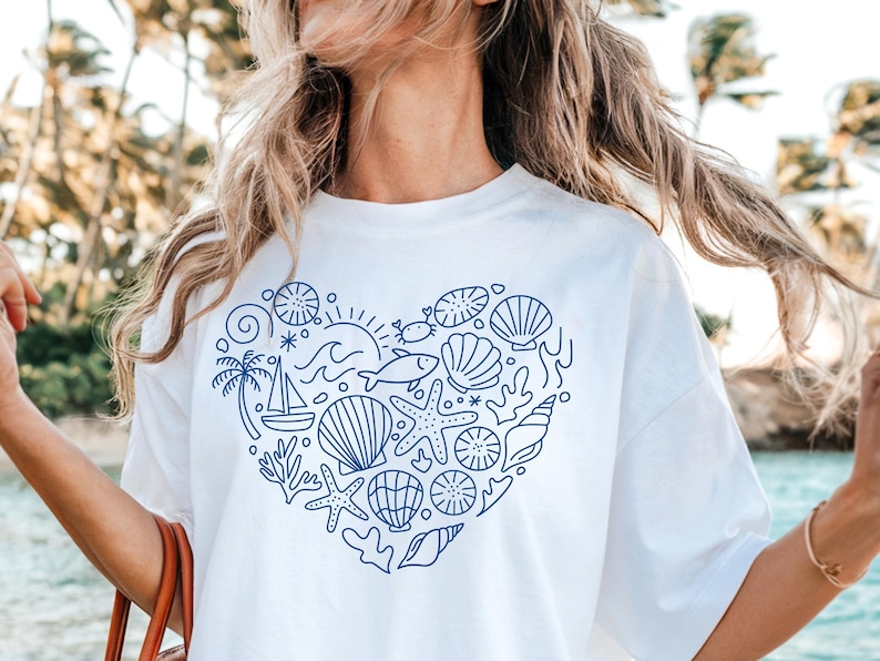 May include: White t-shirt featuring a blue heart-shaped design filled with nautical elements. The design includes seashells, starfish, a sailboat, and a palm tree. The shirt is a casual, everyday wear item.