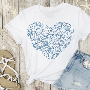 May include: White t-shirt with a blue heart-shaped design filled with nautical elements like seashells, starfish, and a sailboat. The shirt is paired with denim shorts and sandals, suggesting a beach or summer theme.
