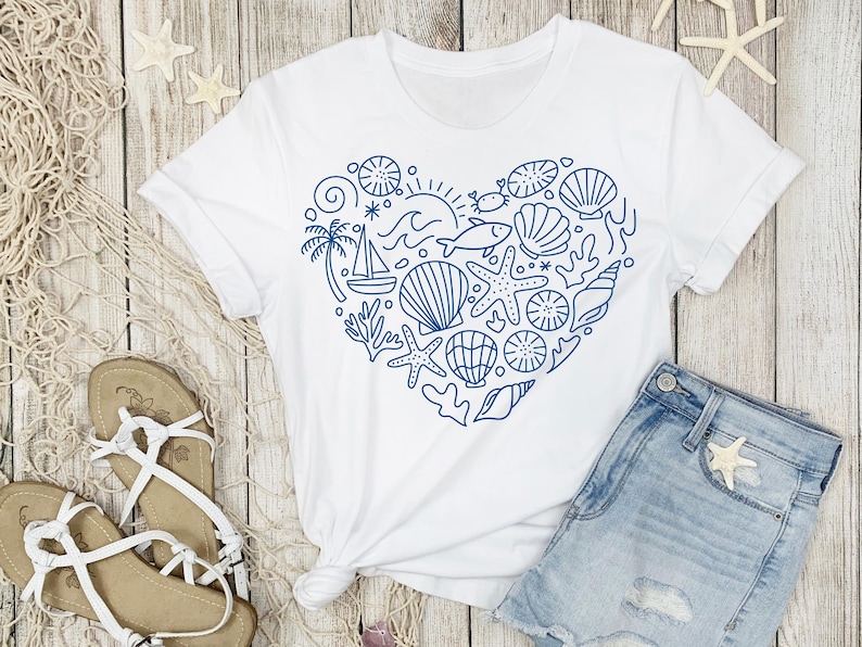 May include: White t-shirt with a blue heart-shaped design filled with nautical elements like seashells, starfish, and a sailboat. The shirt is paired with denim shorts and sandals, suggesting a beach or summer theme.