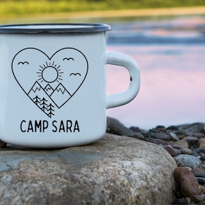 Może przedstawiać: Biały emaliowany kubek do campingu z czarnym konturem serca, g&oacute;r, słońca i drzew. Tekst "CAMP SARA" jest nadrukowany pod sercem.