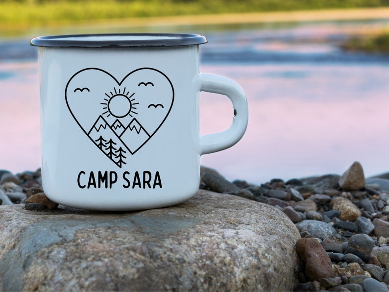 Może przedstawiać: Biały emaliowany kubek do campingu z czarnym konturem serca, g&oacute;r, słońca i drzew. Tekst "CAMP SARA" jest nadrukowany pod sercem.